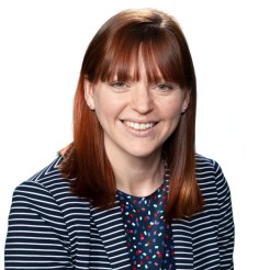 A headshot of Carolyn Nikoloski, a smiling female with red hair wearing a striped navy jacket